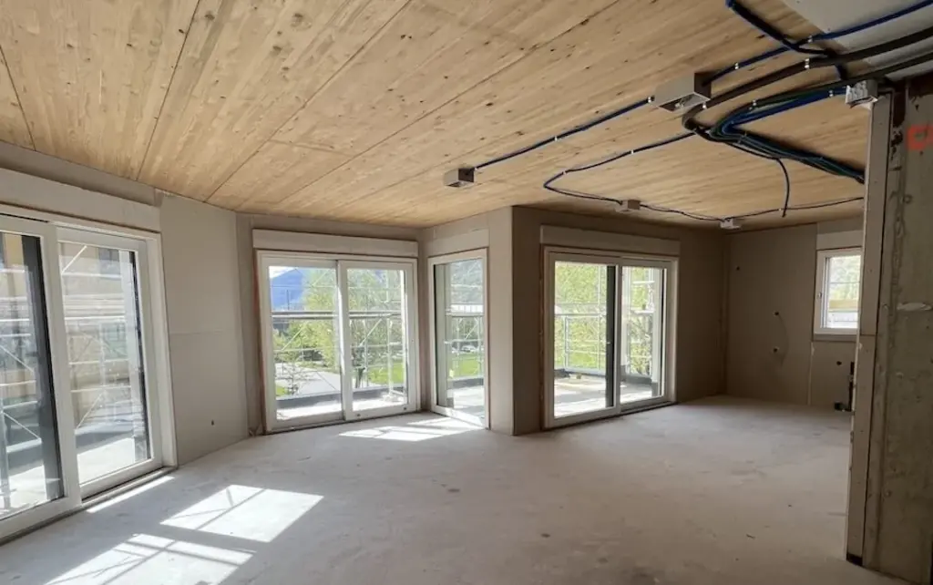 Wohnraum im Rohbau mit Holzdecke, offenen Leitungen und Fensterfront