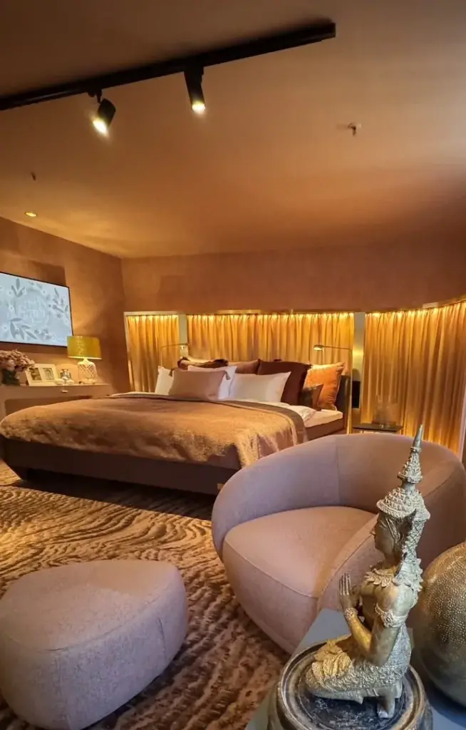 Schlafzimmer mit hinterleuchtetem Vorhang, geschwungenem Sessel und gemustertem Teppich